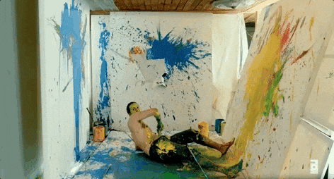 GIF d'un homme torse nu, couvert de peinture, qui glisse sur le sol d'une pièce aux murs blancs éclaboussés de taches de couleurs vives. Il se déplace et roule, créant des projections de peinture aléatoires sur les surfaces autour de lui. La scène évoque une performance artistique chaotique et expressive.