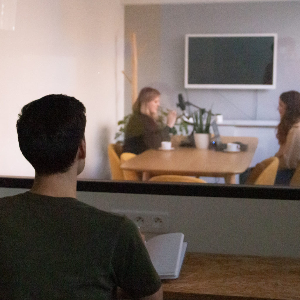 Scène d'observation utilisateur avec une personne au premier plan prenant des notes derrière une vitre, tandis que deux personnes discutent autour d'une table équipée d'un microphone dans une salle de réunion.